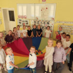 Na zdjęciu widać dzieci z grupy BIEDRONEK. Dzieci stoją w kole na dywanie i trzymają rozłożoną chustę animacyjną.