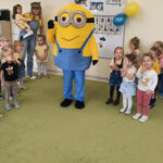 Na zdjęciu widać dzieci z grupy MOTYLKÓW, panią Olę, panią Wiktorię oraz bajkową postać Minionka. Jest to zdjęcie grupowe w sali.