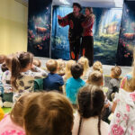 Na zdjęciu widać przedszkolaki siedzące na dywanie i oglądające przedstawienie teatralne.