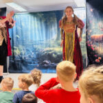 Na zdjęciu widać przedszkolaki siedzące na dywanie i oglądające przedstawienie teatralne oraz dwóch aktorów na scenie.
