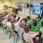 Na zdjęciu widać dzieci z grupy BIEDRONEK, dzieci siedzą na krzesełkach i oglądają na monitorze prezentację edukacyjną.