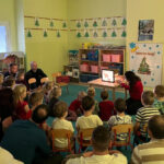 Na zdjęciu widać dzieci z grupy SŁONECZEK, ich rodziców oraz panią Beatę. Pani Beata wykonuje teatrzyk cieni, reszta osób ogląda teatrzyk. Zdjęcie jest robione w sali.