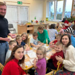 Na zdjęciu widać dzieci z grupy MISIAKÓW i ich rodziców. Wszyscy siedzą przy stole i wykonują pracę plastyczną. Zdjęcie jest zrobione w sali.