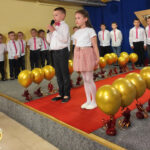 Na zdjęciu widać absolwentów z grupy BIEDRONEK i JEŻYKÓW podczas występu artystycznego. Dzieci stoją na scenie.