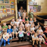 Na zdjęciu widać dzieci z grupy BIEDRONEK, Panią Sandrę oraz Panią Karolinę. Wszyscy siedzą na drewnianych schodach, jest to zdjęcie grupowe w parku atrakcji Jelonkowo.